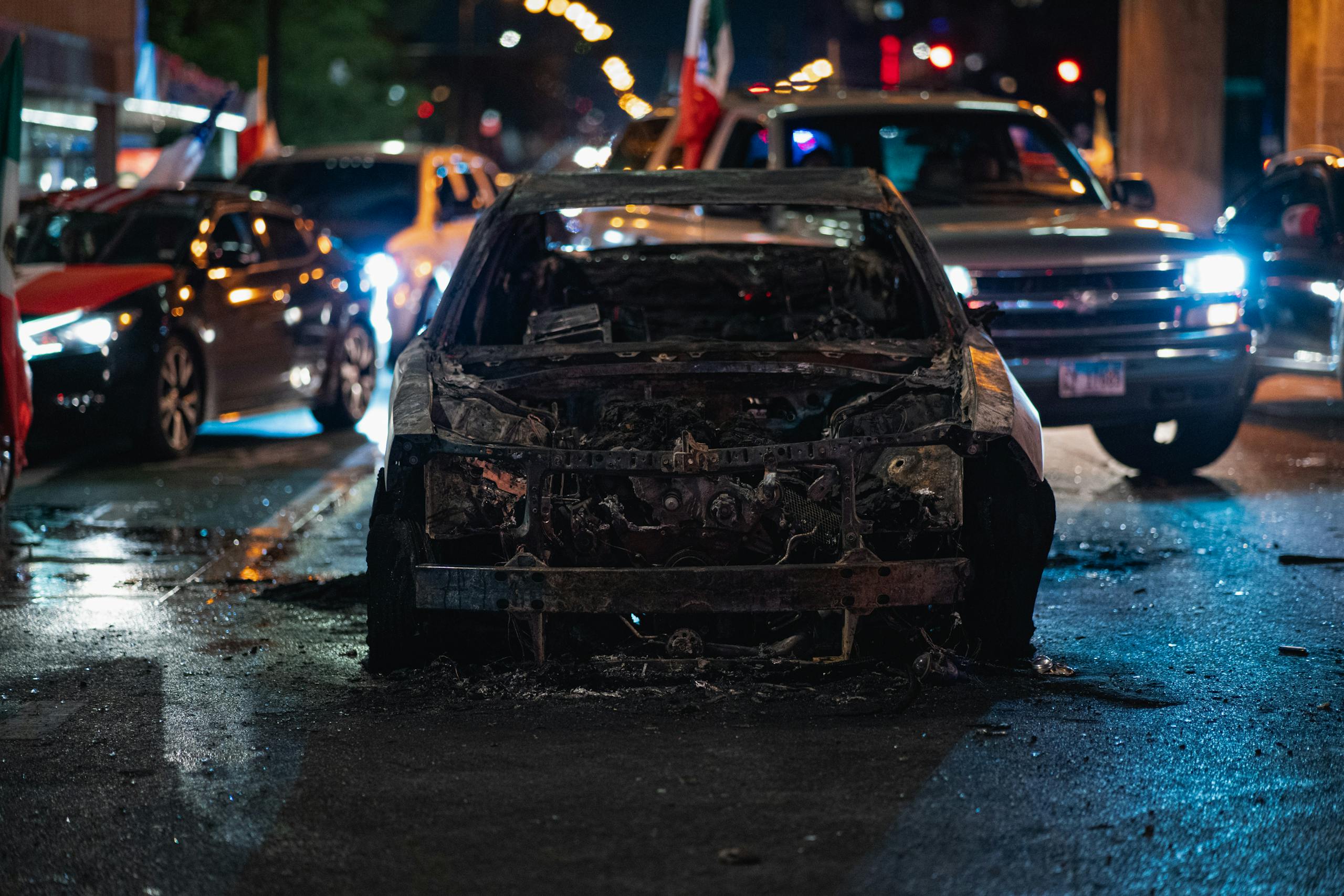 A burnt car is prominently displayed on a city street at night, surrounded by traffic.